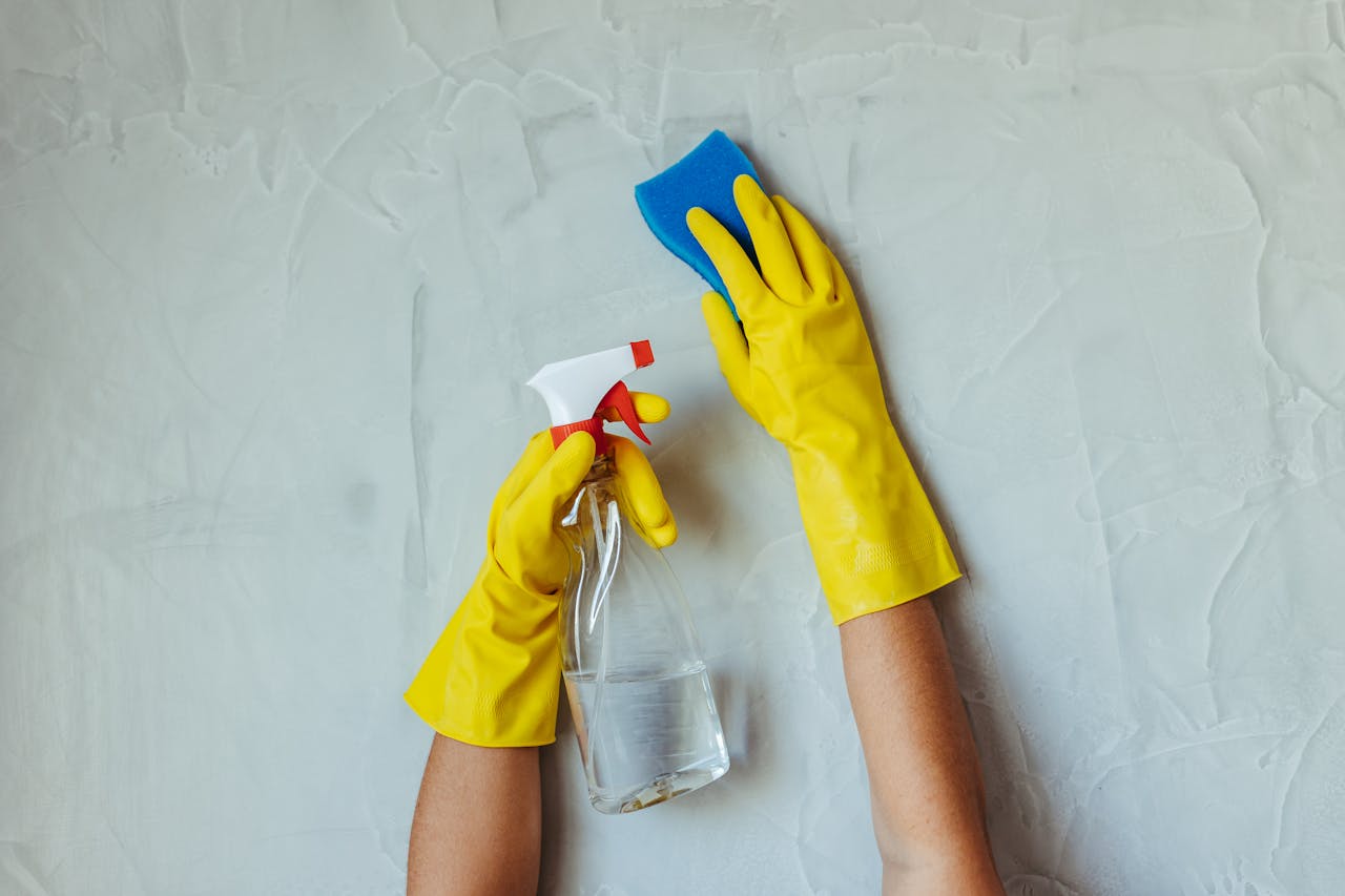 gallery-6 Close-up of hands in yellow gloves cleaning a wall with a spray bottle and sponge.